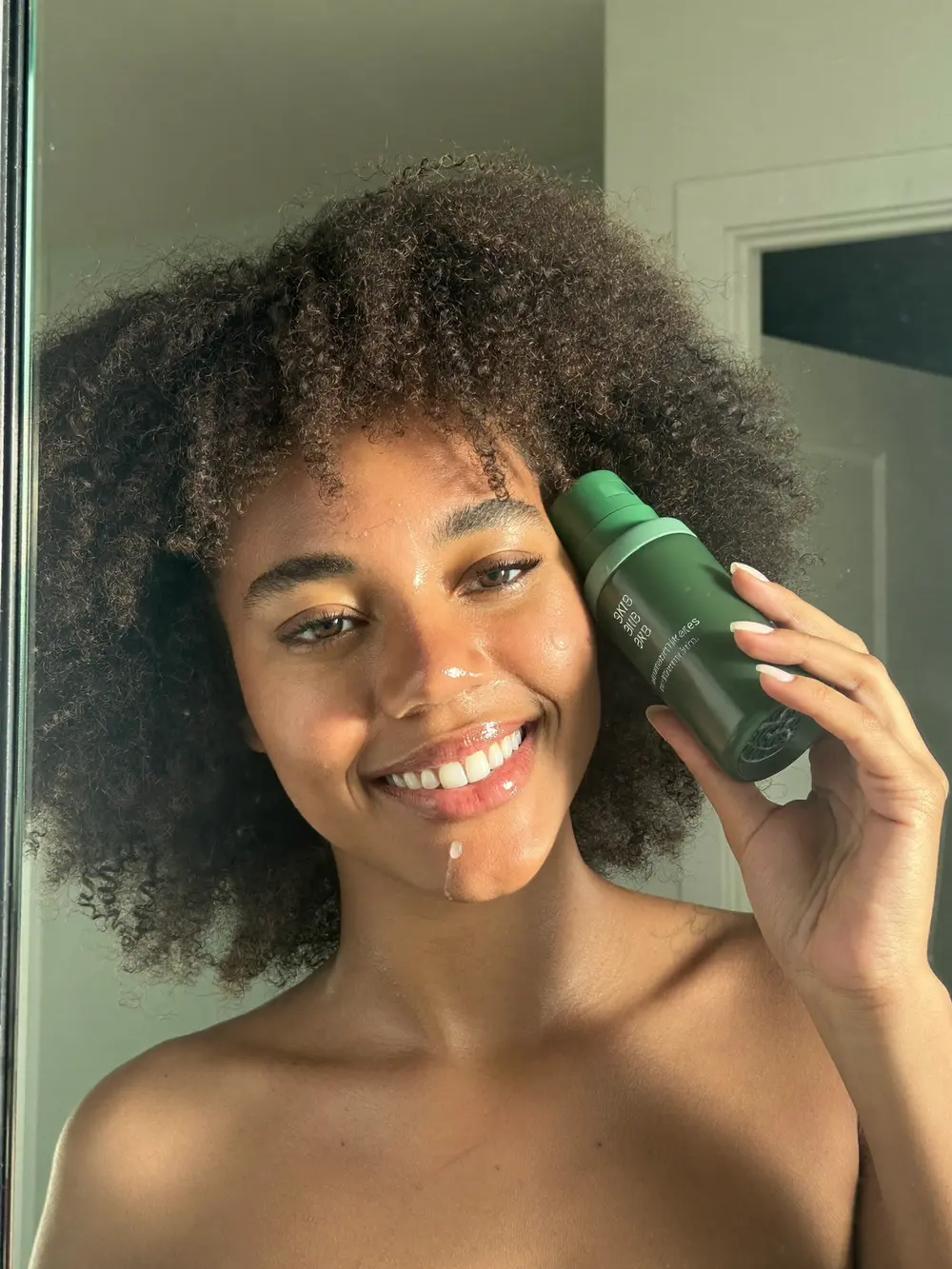 Jovem de cabelo cacheado aplicando sérum no rosto diante do espelho