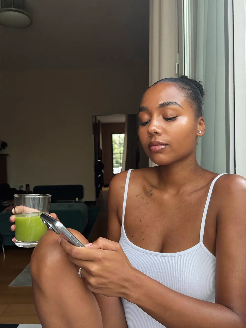 Mulher morena sentada perto da janela lendo no celular, com um copo de água